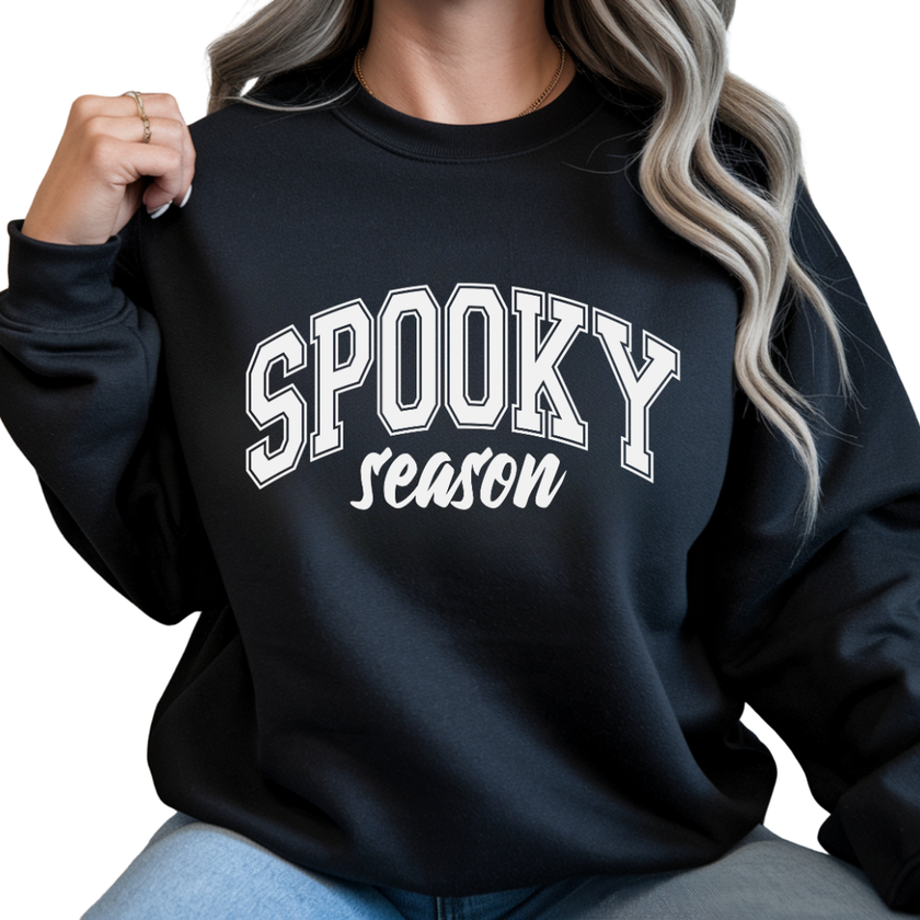 Woman wearing a black Spooky Season graphic sweatshirt featuring large white collegiate Halloween lettering