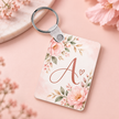 A rectangular acrylic keychain with a silver split ring, decorated with pink peony flowers, green leaves, and a rose gold monogram letter 'A' on a light pink watercolor background, styled on a pink surface with soft floral props.