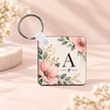 A square personalized luggage tag with a floral design and the letter 'A' monogram, attached to a silver key ring on a soft beige background.