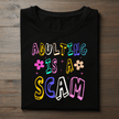 ‘Adulting Is a Scam’ in colorful retro-style font. Ideal for casual wear, birthday gifts, or anyone who loves a good laugh.