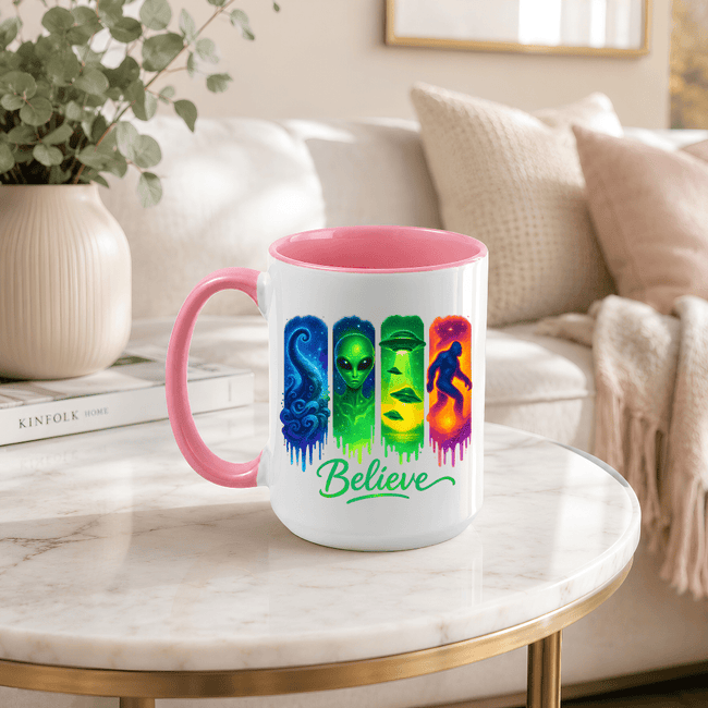 Pink accent Believe alien UFO Bigfoot coffee mug displayed on a table with colourful paranormal themed artwork.