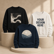 Three crewneck sweatshirts displayed on beige pedestals in a warm studio setting: a black sweatshirt with a gray abstract wave graphic, a cream sweatshirt with the text 'YOUR DESIGN HERE', and a navy sweatshirt with a mountain landscape graphic inside a circle.