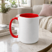 A white ceramic mug with a bright red handle, red interior, and red rim, sitting on a light stone table in a softly blurred living room setting.