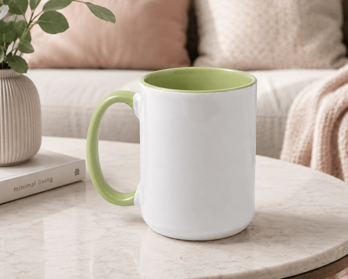 A white ceramic mug with a light green handle and interior, sitting on a round marble table in a cozy living room setting with a sofa and cushions in the background.
