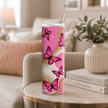 A tall insulated tumbler with a clear lid and metal straw, decorated with pink butterflies and gold glitter on a pink and white background, set on a round marble-top table in a cozy living room.