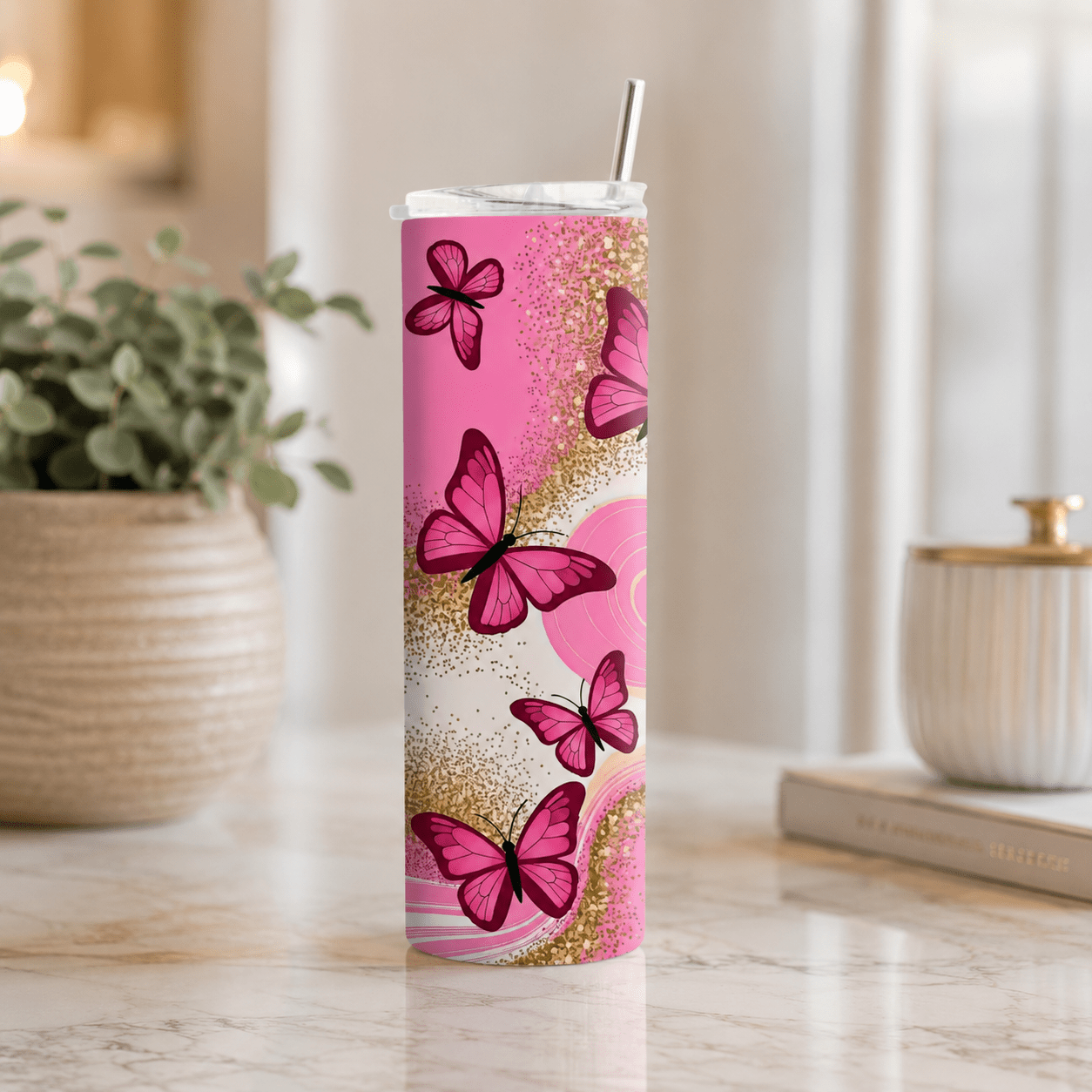 A tall, slim stainless steel tumbler with a clear lid and metal straw, decorated with a pink and gold butterfly pattern, set on a marble countertop in a bright kitchen environment.