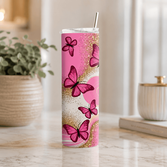A tall, slim stainless steel tumbler with a clear lid and metal straw, decorated with a pink and gold butterfly pattern, set on a marble countertop in a bright kitchen environment.