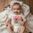 Baby wearing I Steal Hearts dinosaur onesie smiling on soft bedding