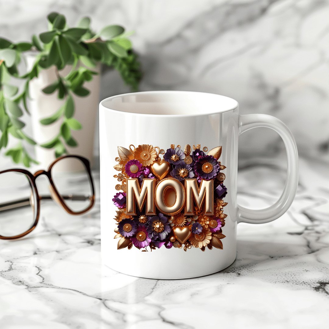 White ceramic MOM mug with gold lettering and floral design placed on marble counter with glasses and plant