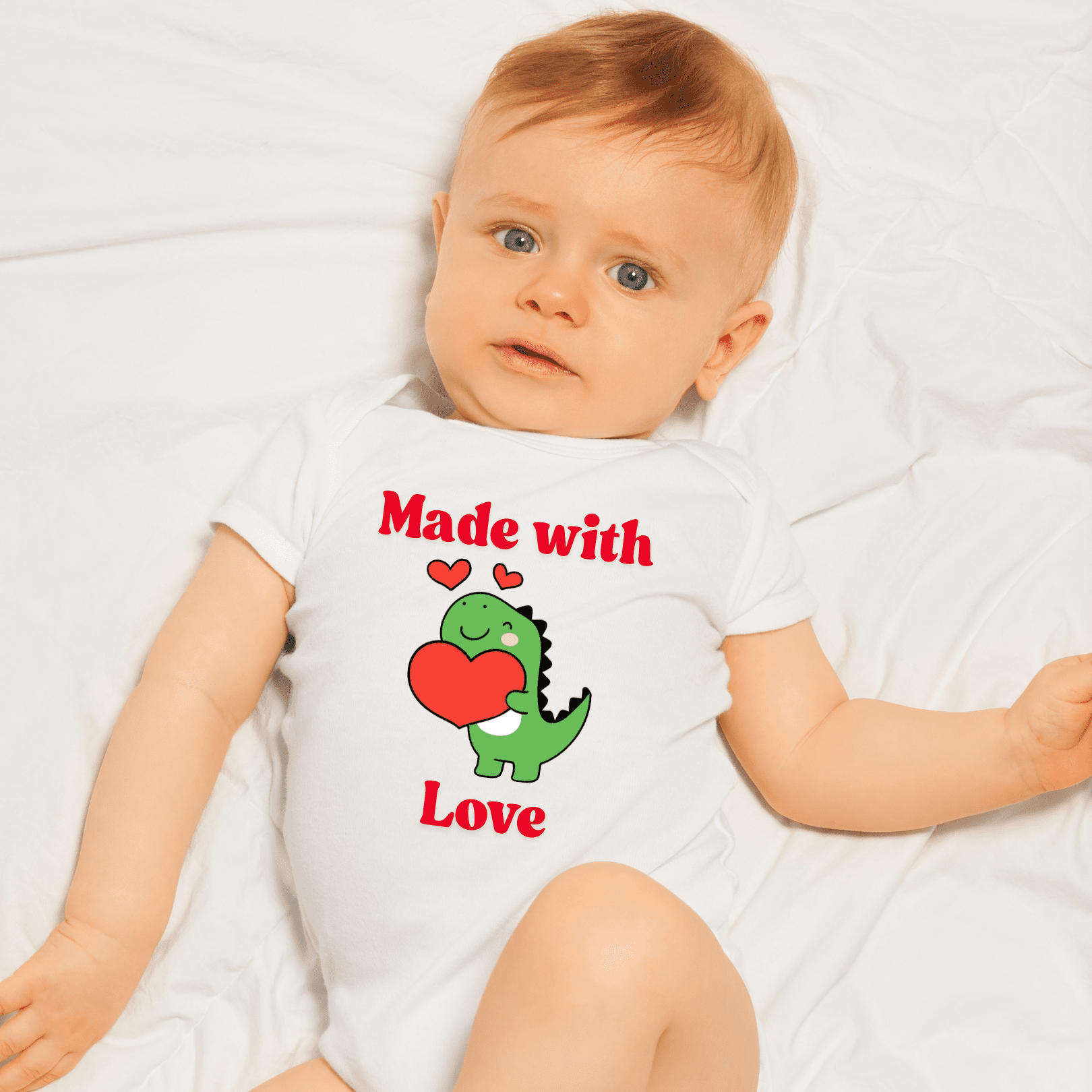 Baby wearing a white onesie with a dinosaur and heart design, laying on a white blanket.