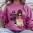 Person wearing a pink sweatshirt with a graphic of a woman holding a coffee cup and text 'Oh Honey I am that Mom' on a wooden background.