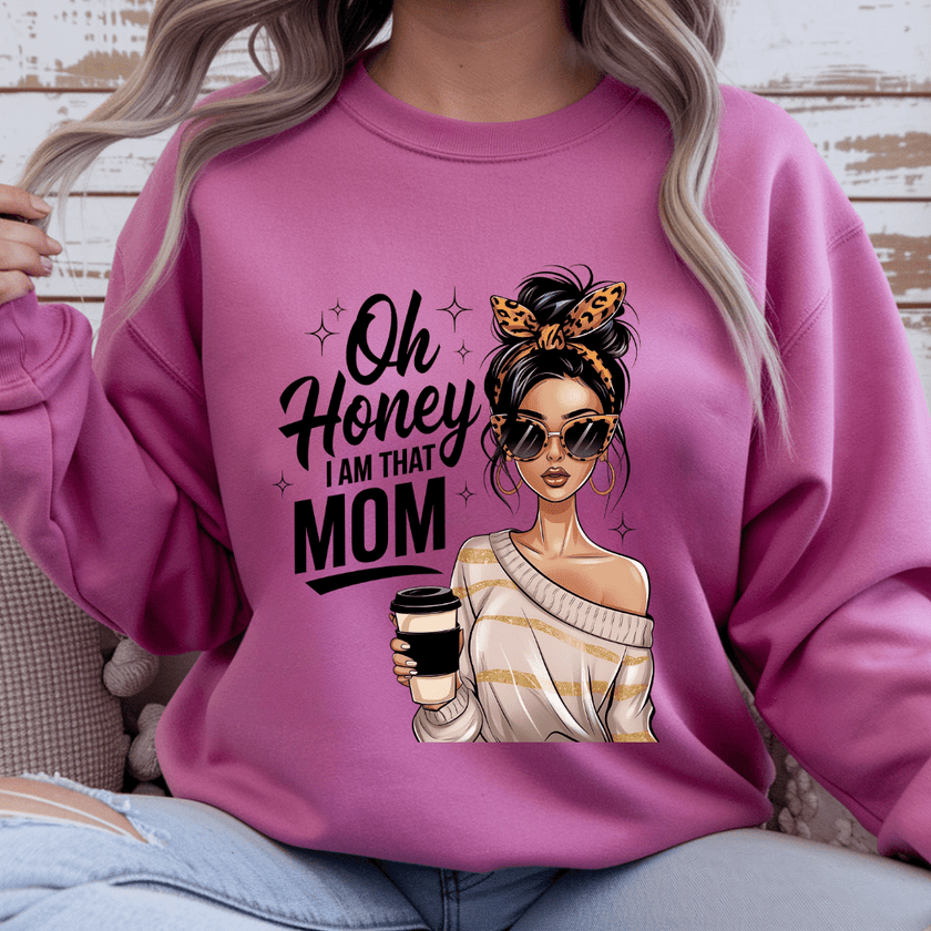 Person wearing a pink sweatshirt with a graphic of a woman holding a coffee cup and text 'Oh Honey I am that Mom' on a wooden background.