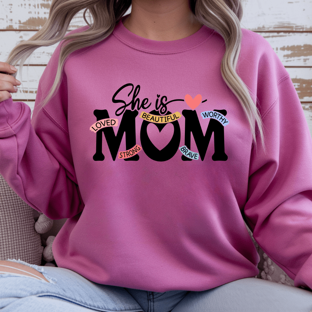 Pink crewneck sweater with “She Is Mom” inspirational graphic featuring words loved strong beautiful worthy brave