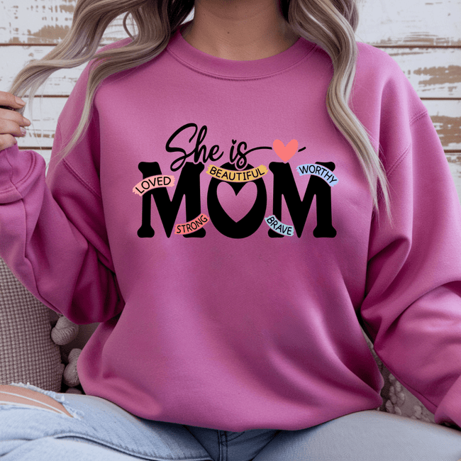 Pink crewneck sweater with “She Is Mom” inspirational graphic featuring words loved strong beautiful worthy brave