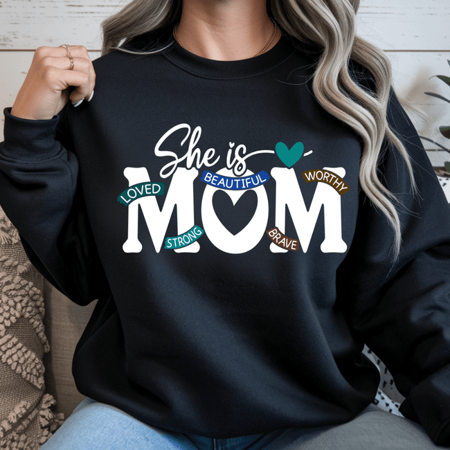 Black sweatshirt with 'She is MOM' text and colorful words on a wooden floor background