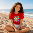 Red youth T shirt worn at the beach featuring the colourful Shine Bright Lil Dumplin rainbow and cute dumpling graphic.