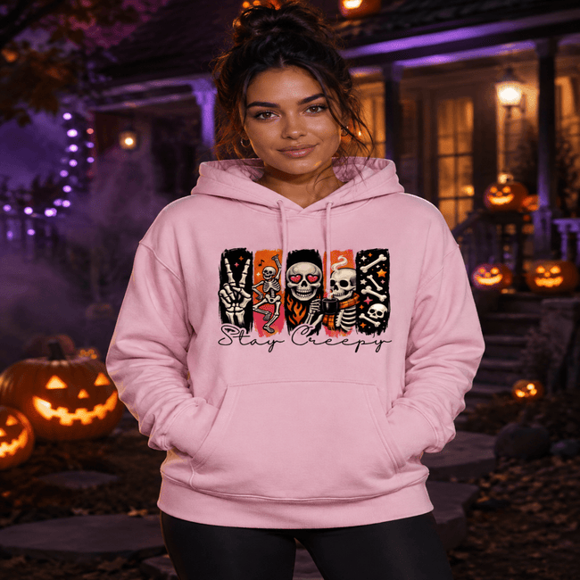 A woman wearing a light pink hoodie with a Halloween-themed graphic print featuring skeletons, skulls, and the text 'Stay Creepy' in front of a spooky house at night with jack-o'-lanterns.