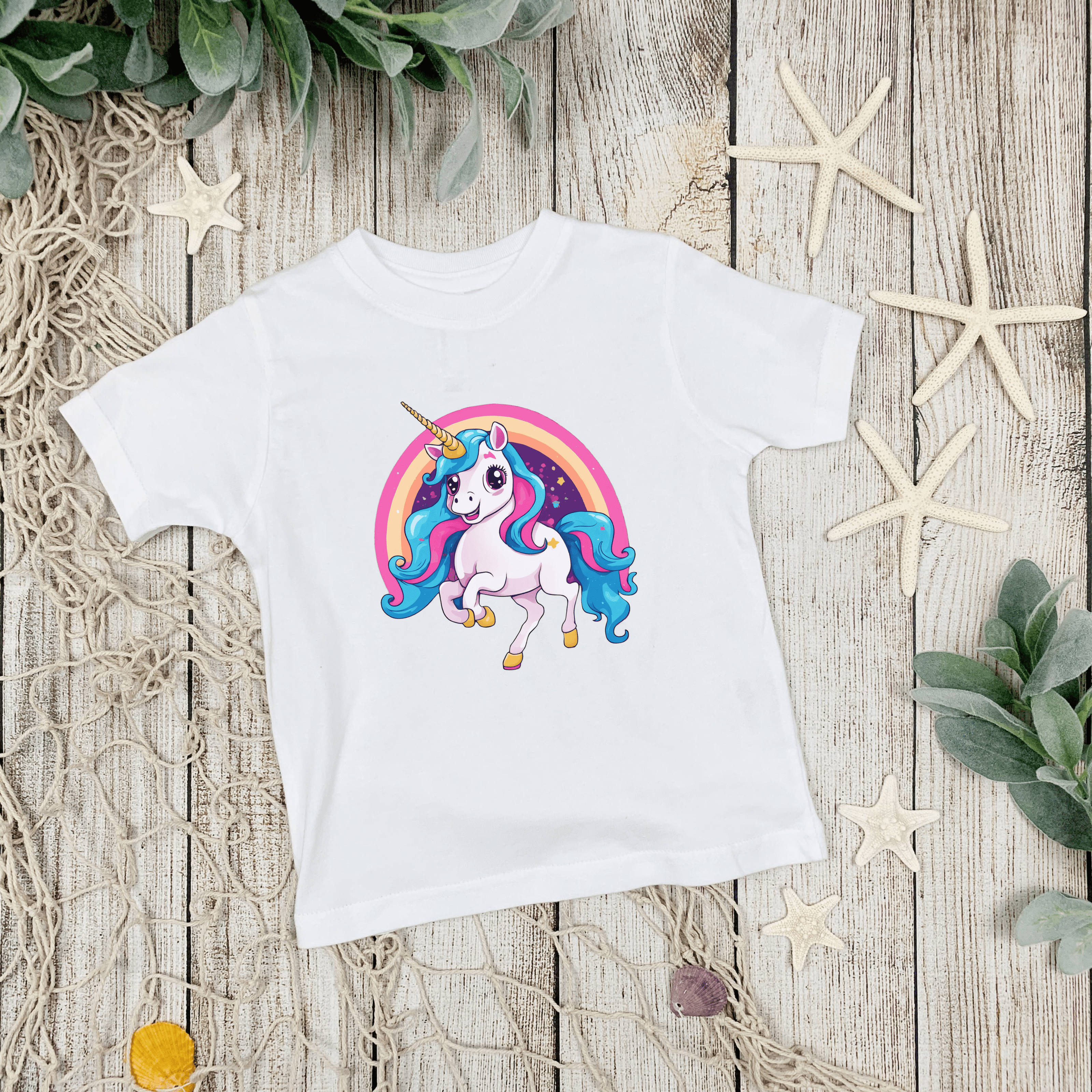 White t-shirt with a colorful unicorn design on a wooden surface with decorative elements.