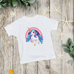 White t-shirt with a colorful unicorn design on a wooden surface with decorative elements.