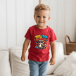 A young child wearing a red graphic t-shirt with a raccoon and the text 'TOO COOL SCHOOL!' printed on the front, standing in a bright living room setting.