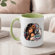 A white ceramic mug with a light green handle and interior, featuring a colorful graphic of a woman with a dreamcatcher headband, flowers, a wolf, and the words 'Wild Heart'. The mug sits on a marble table in a cozy living room setting.