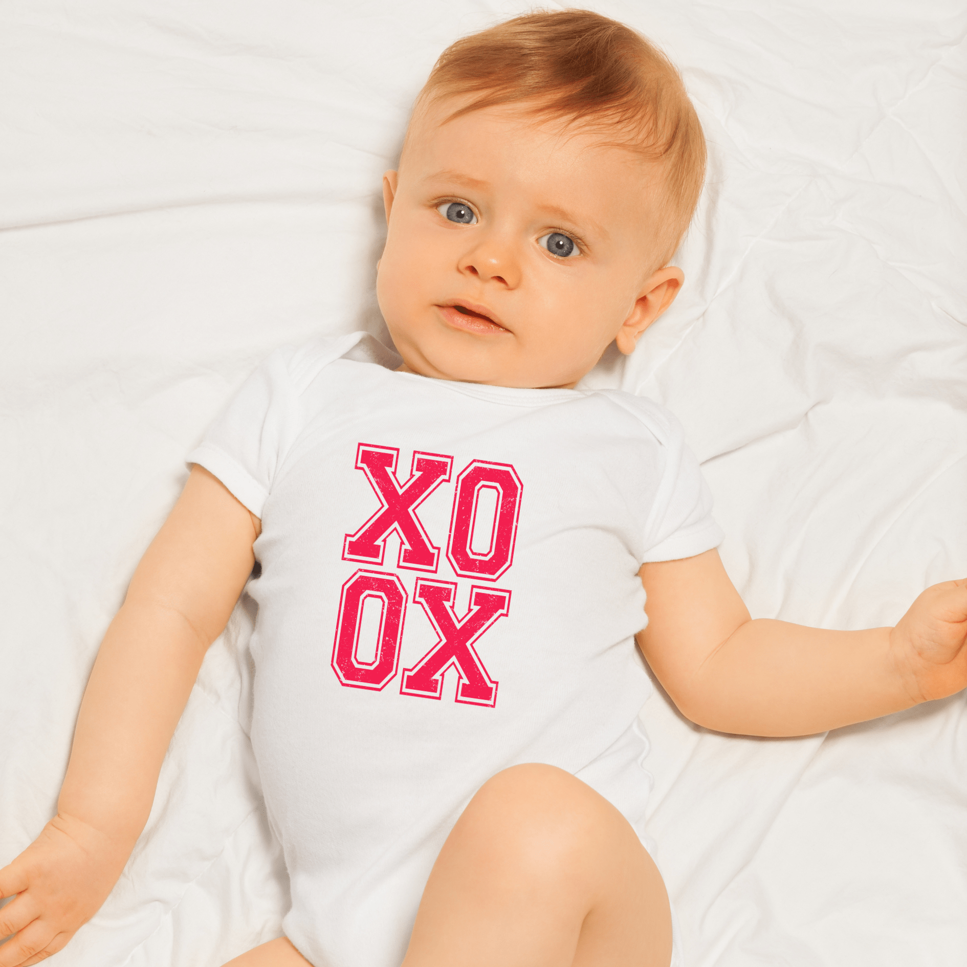 Baby wearing white XO XO onesie with bright pink distressed text lying on white sheet