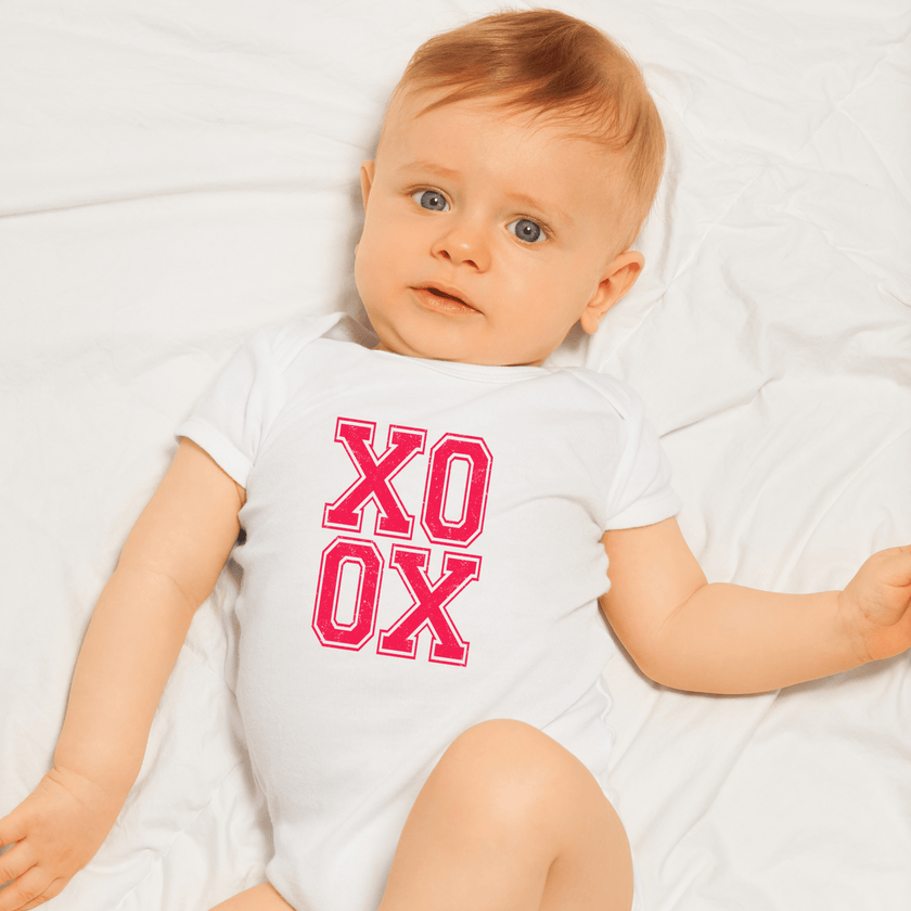 Baby wearing white XO XO onesie with bright pink distressed text lying on white sheet